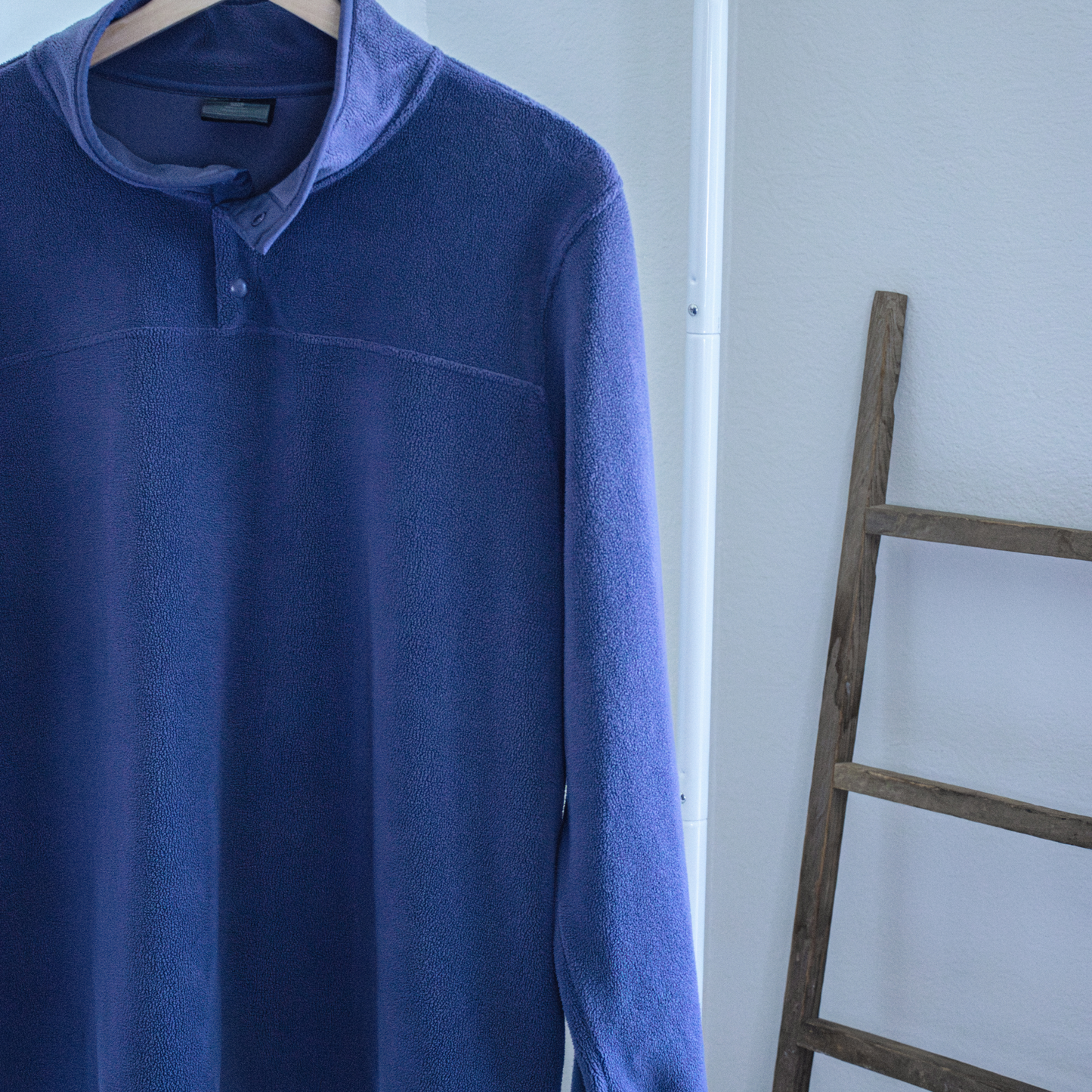 Blue long-sleeve fleece pullover on a hanger against a white wall with a wooden ladder.