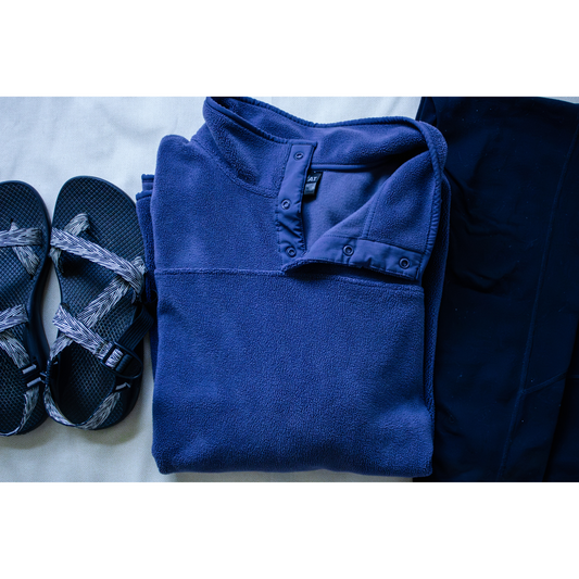 Folded blue fleece sweatshirt next to black Chacos and Sketcher leggings on a light background