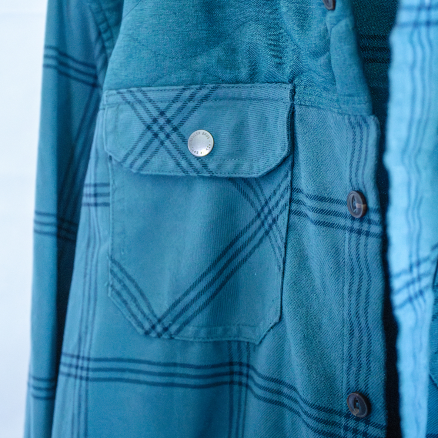 Close-up of a Howler Bros blue plaid shirt with a pocket and buttons on a white background